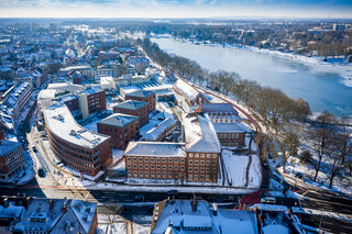 Das Bild zeigt eine Winteransicht einer Stadt mit schneebedeckten Gebäuden und einem gefrorenen Fluss im Vordergrund. Die Architektur ist modern und eindrucksvoll.