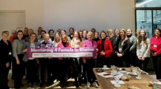 Das Bild zeigt eine Gruppe von Frauen, die zusammenstehen und ein Banner mit der Aufschrift "Mentoring 4 Female in Rheine" halten. Sie wirken freundlich und motiviert.