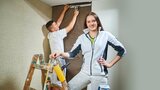 Auf dem Bild sind zwei Personen beim Tapezieren zu sehen. Eine Person lächelt und hält eine Farbrolle, während die andere oben an der Wand arbeitet.