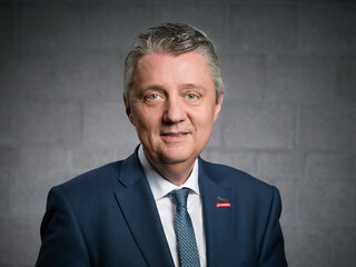 Der Mann trägt einen Anzug und eine Krawatte. Er blickt freundlich in die Kamera, vor einem neutralen, grauen Hintergründe.