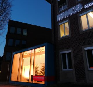 Das Bild zeigt ein modernes Gebäude bei Dämmerung, beleuchtet mit orangefarbenem Licht. Ein Schild mit der Aufschrift "HWK" ist gut sichtbar.