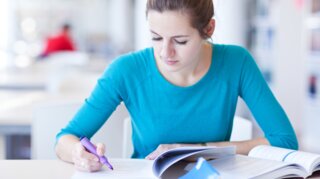 Eine junge Frau sitzt an einem Tisch, liest ein Buch und macht sich Notizen mit einem bunten Textmarker. Im Hintergrund sind Bücherregale sichtbar.