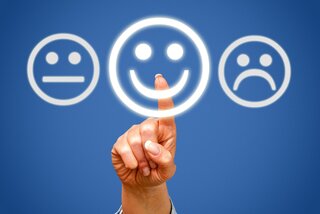 Eine Hand zeigt auf ein leuchtendes Smiley-Gesicht, umgeben von neutralen und traurigen Gesichtern. Symbolisiert Stimmung oder Feedback.