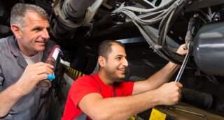 Zwei Mechaniker arbeiten unter einem Fahrzeug. Der eine hält ein Werkzeug und lächelt, während der andere konzentriert eine Schraube löst.