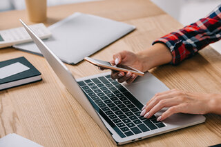 Eine Hand tippt auf der Tastatur eines Laptops, während die andere ein Smartphone hält. Im Hintergrund liegen Notizblätter und eine Tasse.