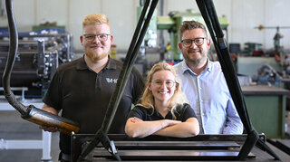 Das Bild zeigt drei Personen in einer Werkstatt. Zwei stehen lächelnd vorne, während der dritte hinter ihnen steht und auf eine Maschine zeigt.