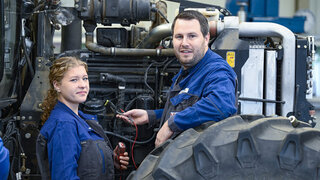 Das Bild zeigt zwei Mechaniker, eine Frau und einen Mann, in Arbeitskleidung, die an einem Traktor arbeiten. Sie scheinen konzentriert und professionell.
