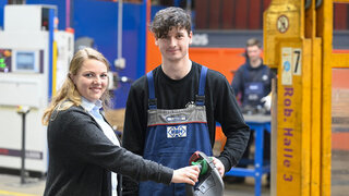 Das Bild zeigt zwei Jugendliche in einer Werkstatt. Die Frau lächelt und hält ein Werkzeug, während der Mann daneben steht, ebenfalls lächelnd.