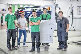 Die Gruppe von fünf Personen steht in einer Werkstatt. Sie tragen Arbeitskleidung und lächeln, während sie an einem Fahrzeug arbeiten.