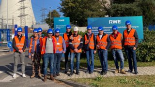 Eine Gruppe von Personen in Sicherheitswesten und Helmen steht vor einem RWE-Werk mit Kühltürmen im Hintergrund. Sonnenlicht beleuchtet die Szene.