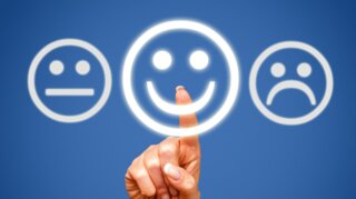 Eine Hand zeigt auf ein leuchtendes, lächelndes Smiley zwischen einem neutralen und einem traurigen Gesicht. Symbol für Emotionen oder Umfragen.