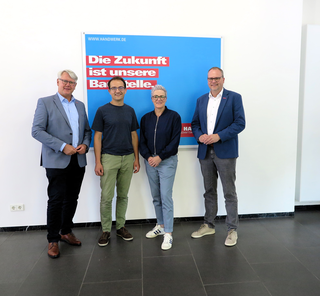 Das Bild zeigt vier Personen in einem modernen Raum vor einem Plakat mit der Aufschrift „Die Zukunft ist unsere Baustelle“. Sie wirken freundlich und harmonisch.