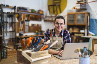 Eine Handwerkerin in einem Werkraum sitzt lächelnd an einem Tisch mit einem Laptop. Werkzeuge und Holzmaterialien sind um sie herum angeordnet.