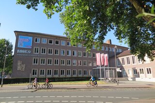 Das Bild zeigt die Handwerkskammer Münster an einem sonnigen Tag. Im Vordergrund fahren Radfahrer vorbei, vor einem modernen Gebäude mit Fenstern und Fahnen.