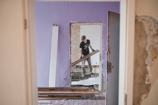 Ein Mann renoviert einen Raum, entfernt Holzplatten und zeigt den Fortschritt der Umbauarbeiten in einem stark beschädigten Raum.