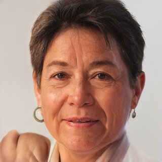 Das Bild zeigt eine Frau mit kurzen, grauen Haaren und einem freundlichen Ausdruck. Sie trägt Ohrringe und hat eine entspannte Haltung.
