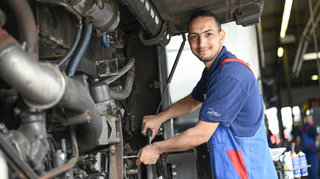 Ein Mechaniker arbeitet an einem Motor. Er trägt eine blaue Arbeitskleidung und lächelt, während er ein Werkzeug hält.