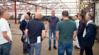 Eine Gruppe von Männern steht in einer Halle und hört einem Vortragenden zu. Die Atmosphäre scheint informell und aufmerksam.