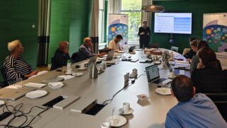 Das Bild zeigt eine Besprechung in einem modernen Konferenzraum. Teilnehmer hören einem Präsentierenden zu, während Notizen und Laptops auf dem Tisch liegen.